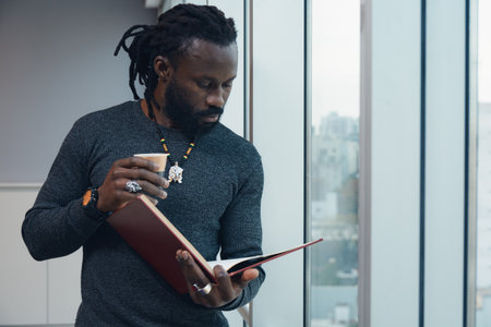 Young Man with dreadlocks and beard attentively reading a book standing by the window in a Coworking while holding a cup of coffee, the man enjoys his coffee and book in a comfortable settingの写真素材