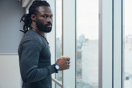 Man With dreadlocks looking at camera is holding a cup of coffee in front of a window in the office. Concept of relaxation and contemplation, as the man enjoys his coffee while take a breakの写真素材