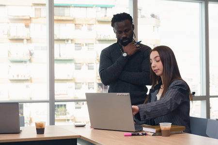 An Adult Man with dreadlocks is standing watching what a Woman is pointing at on the laptop inside...