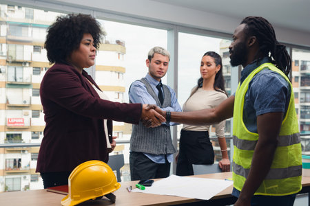 Engineer Man and contracting Woman shaking hands after meeting to close contract, they are inside office around table with documents on it, there are two people in background witnessing deal.の写真素材