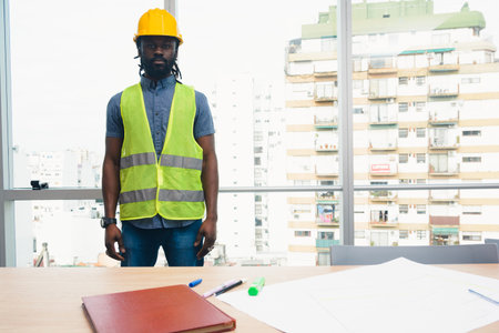 Portrait of Adult Man of African ethnicity engineer uniformed with helmet and vest is standing in...