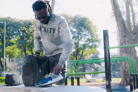 Young African Man standing in the park taking out his running shoes from his backpack for training, he is getting ready for training in the parkの写真素材