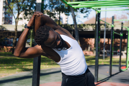 Black African Man with dreadlocks, stands in the park demonstrating how to stretch shoulders and arms correctly, he brings his left arm behind his head and puts pressure on it to stretchの写真素材