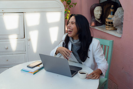 Young Latina woman of Argentine ethnicity, is at home studying and watching funny videos laughing out loud, is sitting and has a laptop and books on the table, people concept, copy spaceの写真素材