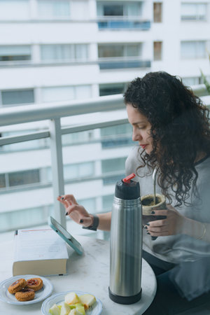 Woman sitting on the balcony making a transaction on the phone while having a quiet breakfast in her apartment, she has a book and food on the table, she is drinking mate, vertical image.の写真素材