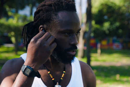 Close-up of a black man with dreadlocks and a beard in the park looking to the side, he is putting on his headphones to exercise listening to music, he is wearing a watch and a white t-shirt, copy space.の写真素材