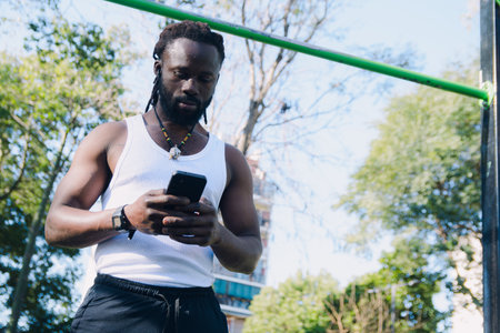 black ethnic man, with dreadlocks and beard, wearing white t-shirt and black pants, standing in calisthenics park texting on phone while resting, copy space, technology concept.の写真素材