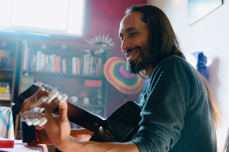 happy long hair bearded Man at home smiling playing guitar, he is learning music, he plays and sees...