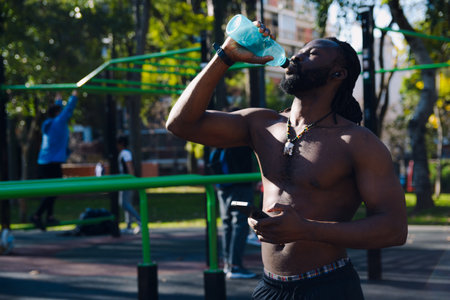Young black sporty man shirtless standing drinking water in calisthenics park, he wears dreadlocks and beard, he is hydrating himself during break from workout routines.の写真素材