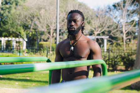 Young African man with dreadlocks and no shirt is training in the calisthenics park, he is standing...