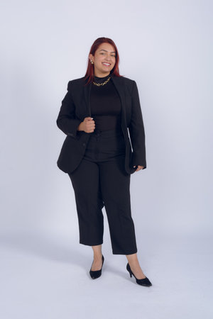 Red haired Colombian entrepreneur woman, confident and elegant, wearing a black business suit, standing posing smiling and looking at the camera, studio photo on white background.の写真素材