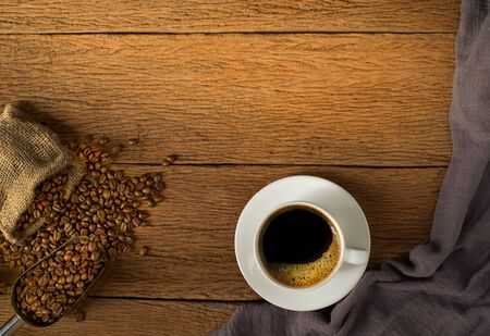 Top view above of Black hot coffee for morning with milk foam for morning menu in white ceramic cup with coffee beans roasted in burlap sack bag on wood table background. Flat lay with copy space.の写真素材