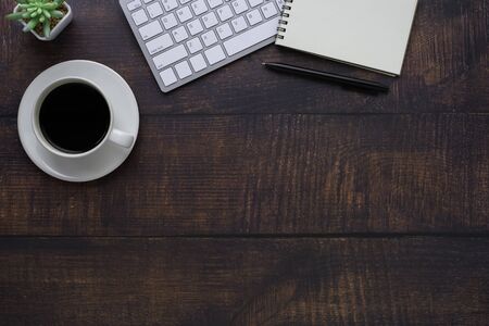 Top view above of Dark wooden office desk table with keyboard, notebook and coffee cup with...