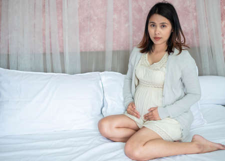 Beautiful asian woman pregnant happy in beige maternity dress sitting on bed at home. Mom is touching and caressing her belly with hands . Motherhood, pregnancy, gynecology and health care concept.の写真素材