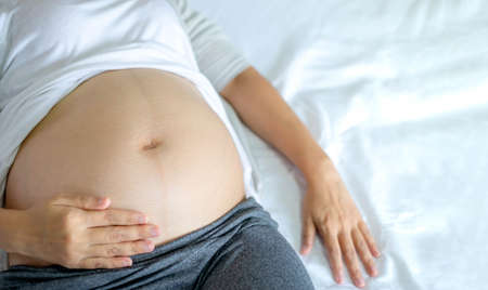 Image of A woman pregnant reclining on bed and touching or caressing her belly with hands. Motherhood, pregnancy, gynecology and health care concept.の写真素材
