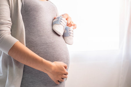 A woman pregnant in gray maternity dress standing beside the window with baby shoes. Mom is touching...
