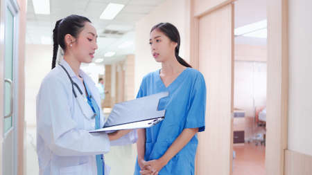 Professional, Asian young female medical doctor, staff in uniform standing in hospital corridor. Two colleague workers discuss, work together with medical report clipboard on clinic hallway.の写真素材