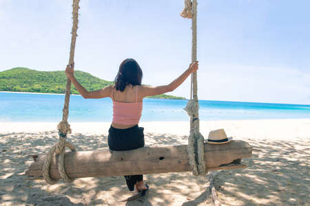 Back Beautiful young Asian women with tan skin wearing pink tank black pants sitting on timber swing. beach and seaside fresh sky background. Holiday, Vacation, Chilling, Relax concept. Alone Concept.の写真素材