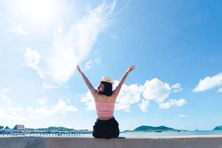 Back of asian woman tan skin wearing pink tank top and straw hat sitting on mortar with standing arms outstretched on sky. looking into the sea. Summer travel. Relaxing, Holiday and tropical relax concept.の写真素材