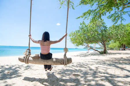 Back Beautiful young Asian women with tan skin wearing pink tank black pants sitting on timber swing. beach and seaside fresh sky background. Holiday, Vacation, Chilling, Relax concept. Alone Concept.の写真素材