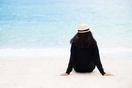 Back of asian woman tan skin wearing black shirt and straw hat sitting on the sand at beach and looking to the sea. Summer travel. Relax, Holiday and tropical relax concept. Alone people concept.の写真素材