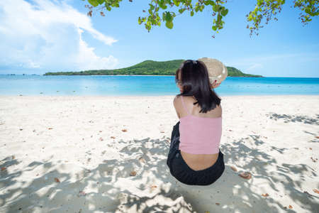 Back of asian woman tan skin wearing pink tank top and straw hat sitting on beach under tree. Summer travel. Relax, Holiday and tropical relax concept. alone concept.の写真素材