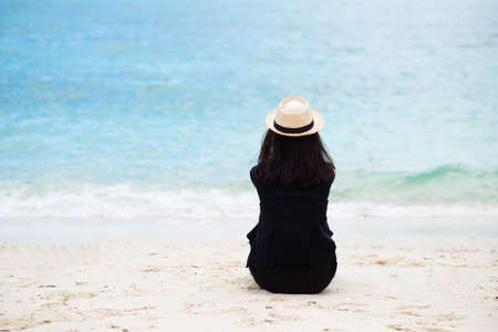 Back of asian woman tan skin wearing black shirt and straw hat sitting on the sand at beach and looking to the sea. Summer travel. Relax, Holiday and tropical relax concept. Alone people concept.の写真素材
