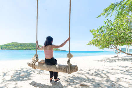 Back Beautiful young Asian women with tan skin wearing pink tank black pants sitting on timber swing. beach and seaside fresh sky background. Holiday, Vacation, Chilling, Relax concept. Alone Concept.の写真素材
