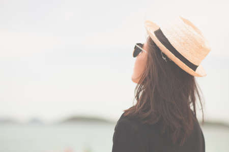Vintage tone medium shot woman tan skin wearing black shirt with straw hat and sunglasses. looking into sea. on sea background. Summer travel. Relaxing, Holiday and tropical. Alone concept.の写真素材