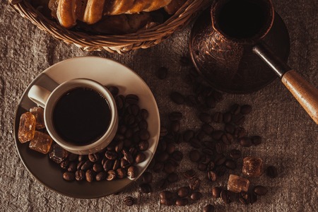 A cup of coffee on textile with coffee beans, dark candy sugar, pots, basket and cakeの写真素材