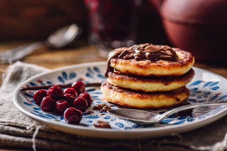 Stack of Quark Pancakes with Hazelnut Paste, Frozen Cherry and Vanilla Pod. Tea Pot with Spoons and Glass of Berries on Background.の写真素材