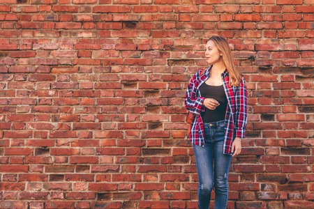 Portrait of young woman in checkered shirt and blue jeans standing against red brick wallの写真素材