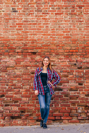 Portrait of young woman in checkered shirt and blue jeans standing against red brick wallの写真素材