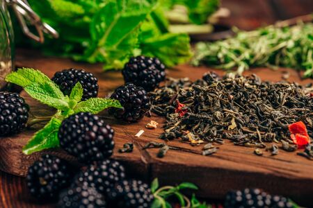 Heap of Dry Green Tea and Fresh Blackberries on Wooden Cutting Board. Bundles of Mint and Thyme...