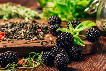 Heap of Dry Green Tea and Fresh Blackberries on Wooden Cutting Board. Bundles of Mint and Thyme...