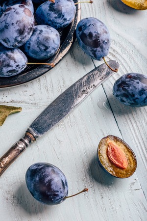 Ripe plums with sliced fruits, leaves and vintage knife over light wooden surface. View from aboveの写真素材