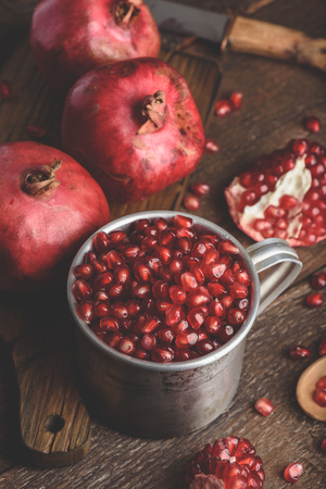 Rustic metal mug full of pomegranate seeds. Whole fruits and pomegranate pieces on dark wooden table.の写真素材
