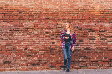 Portrait of young woman in checkered shirt and blue jeans standing against red brick wallの写真素材