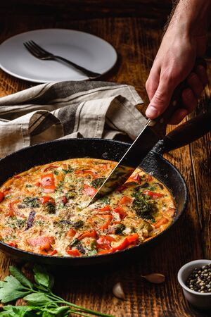 Hand with knife slices vegetable frittata with broccoli, red bell pepper and red onion in cast iron panの写真素材