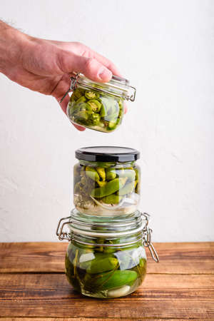 Male Hand Holding One of Three Glass Jars of Freshly Pickled Jalapeno Peppers with Herbs and Garlic. Copy spaceの写真素材