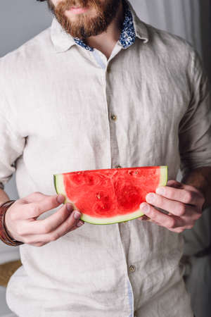 Bearded man in linen shirt holds in his hands slice of ripe red watermelonの写真素材