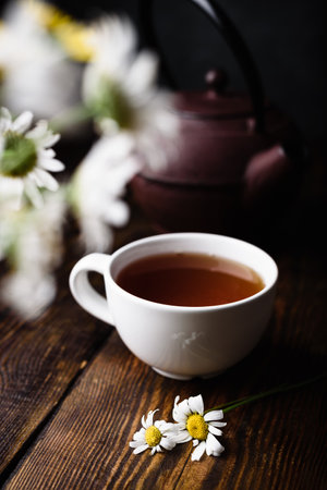 White cup of tea and chamomile flowers on wooden table. Blurred bouquet on foregroundの写真素材