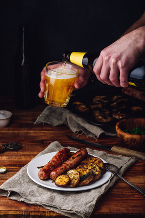 Pork Sausages Baked with Eggplant, Leek and Herbs on White Plate. Man Pouring Beer from Bottle into a Glass.の写真素材
