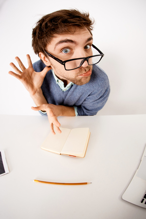Funny and crazy handsome young businessman in glasses sitting at the office desk on white background. Looking at camera. Wide angle.の写真素材