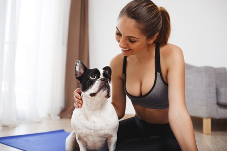 Beautiful fitness girl sitting on yoga-mat on floor with dogの写真素材