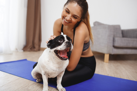 Beautiful fitness girl sitting on yoga-mat on floor with dogの写真素材