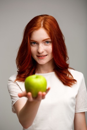 Portrait of young beautiful ginger girl holding green apple, smiling, looking at camera over gray background.の写真素材