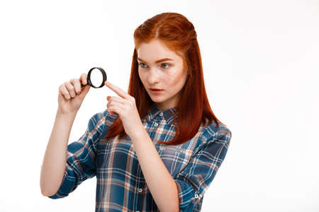 Portrait of young beautiful ginger girl holding magnifier over white background.の写真素材