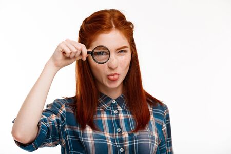 Portrait of young beautiful ginger girl with magnifier looking at camera, showing tongue over white background.の写真素材