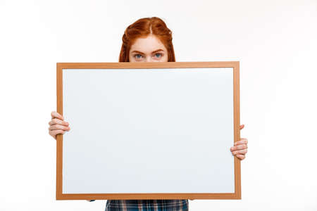 Portrait of young beautiful ginger girl hiding face with whiteboard, looking at camera over white background. Copy space.の写真素材